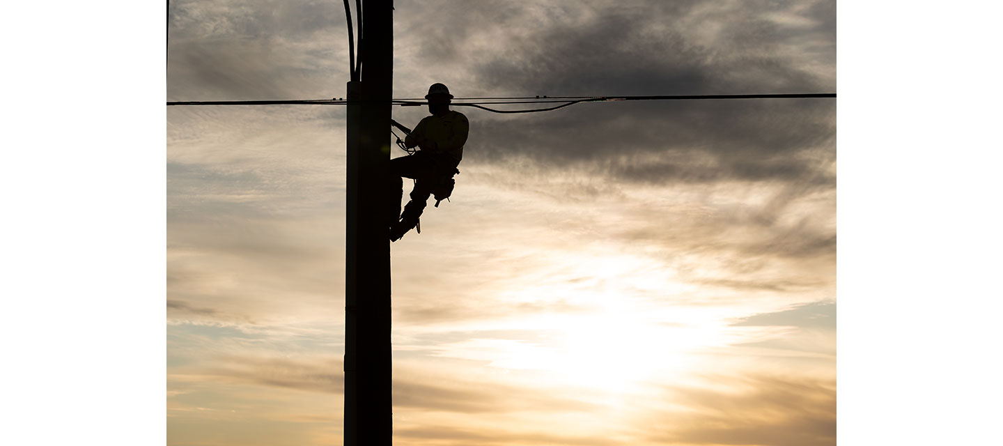 Silhouette of a lineman climbing a utility pole at sunset, secured with safety gear against a cloudy sky.